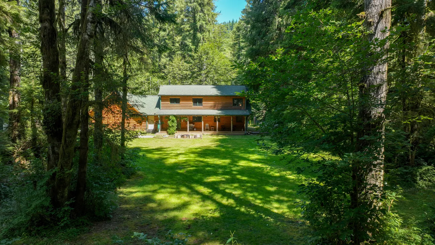 A two-story log cabin nestled in a dense forest with a large, sunny grassy lawn in the foreground.