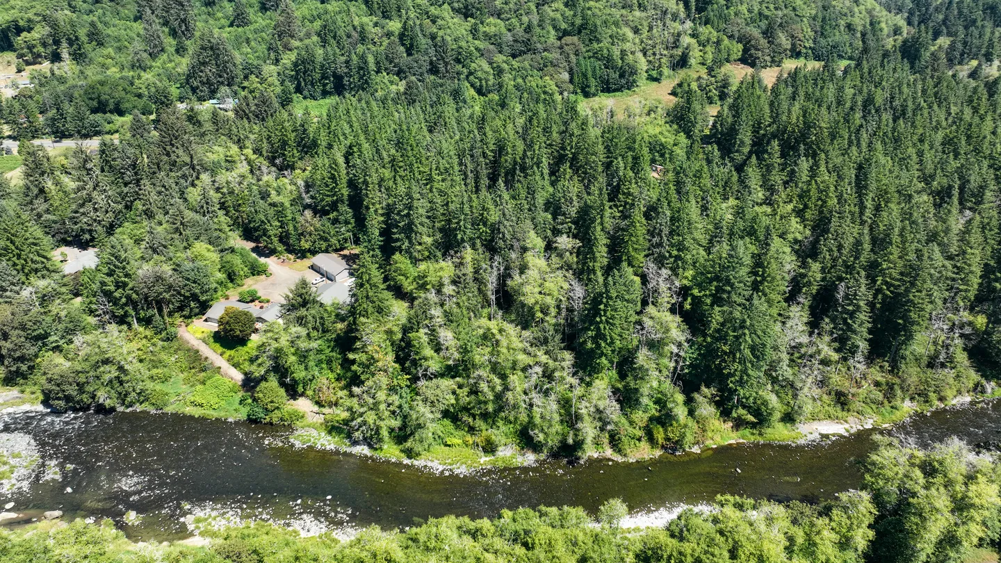 Aerial view of a river winding past dense evergreen forest with house rooftops nestled in the trees.