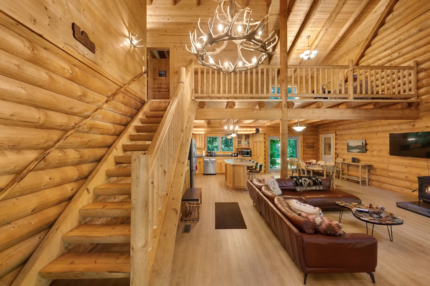 Open-concept log cabin interior with wooden stairs leading to a loft, a living area with a leather sectional, an antler chandelier, and a kitchen in the background.