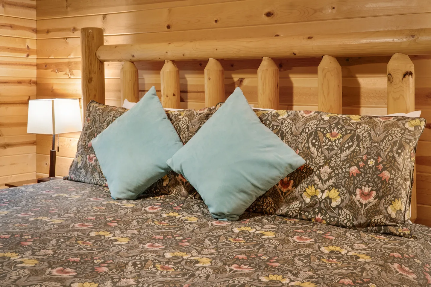 Close-up of a bed with a log headboard, floral patterned bedding, two light blue accent pillows, and a bedside lamp against wood-paneled walls.