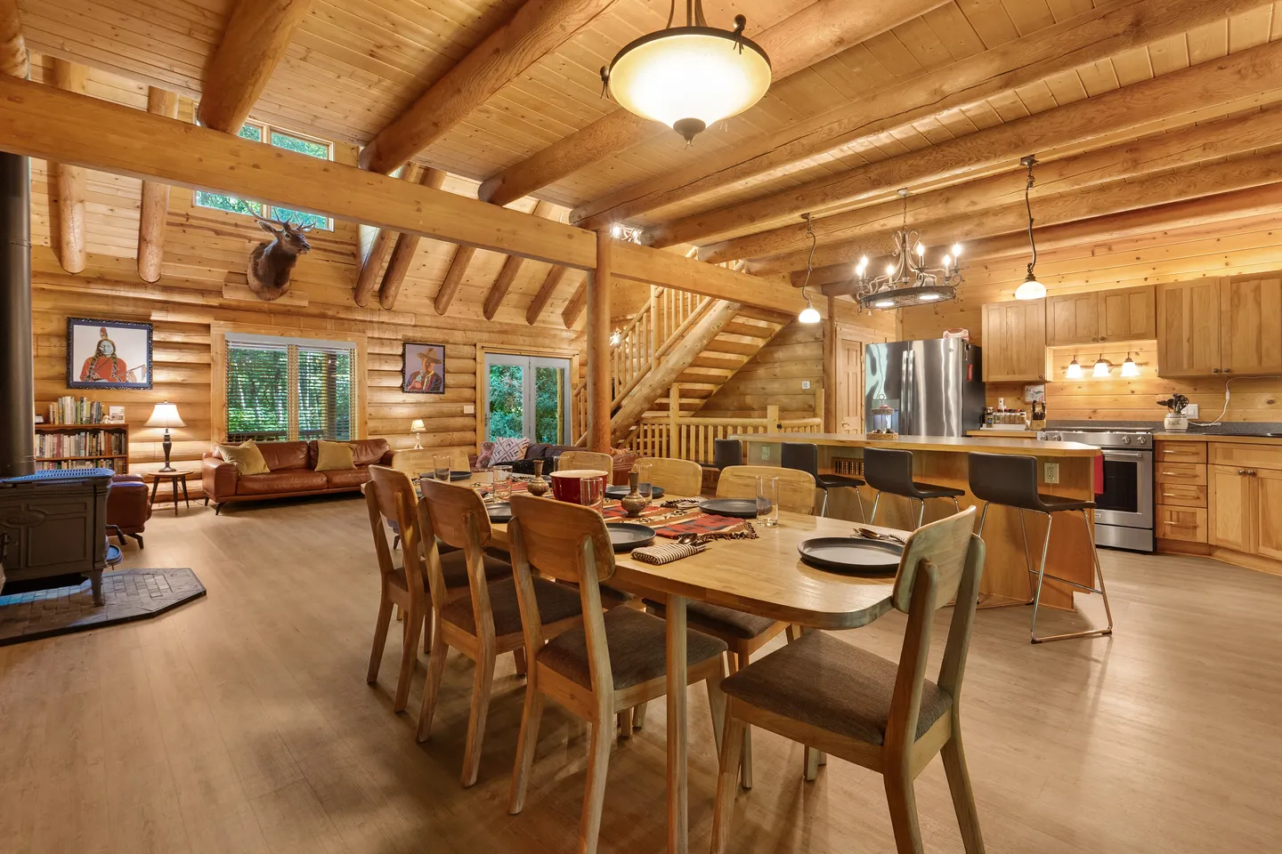 Open-concept log cabin interior showing a dining table with chairs, an adjacent kitchen, and a living area in the background.