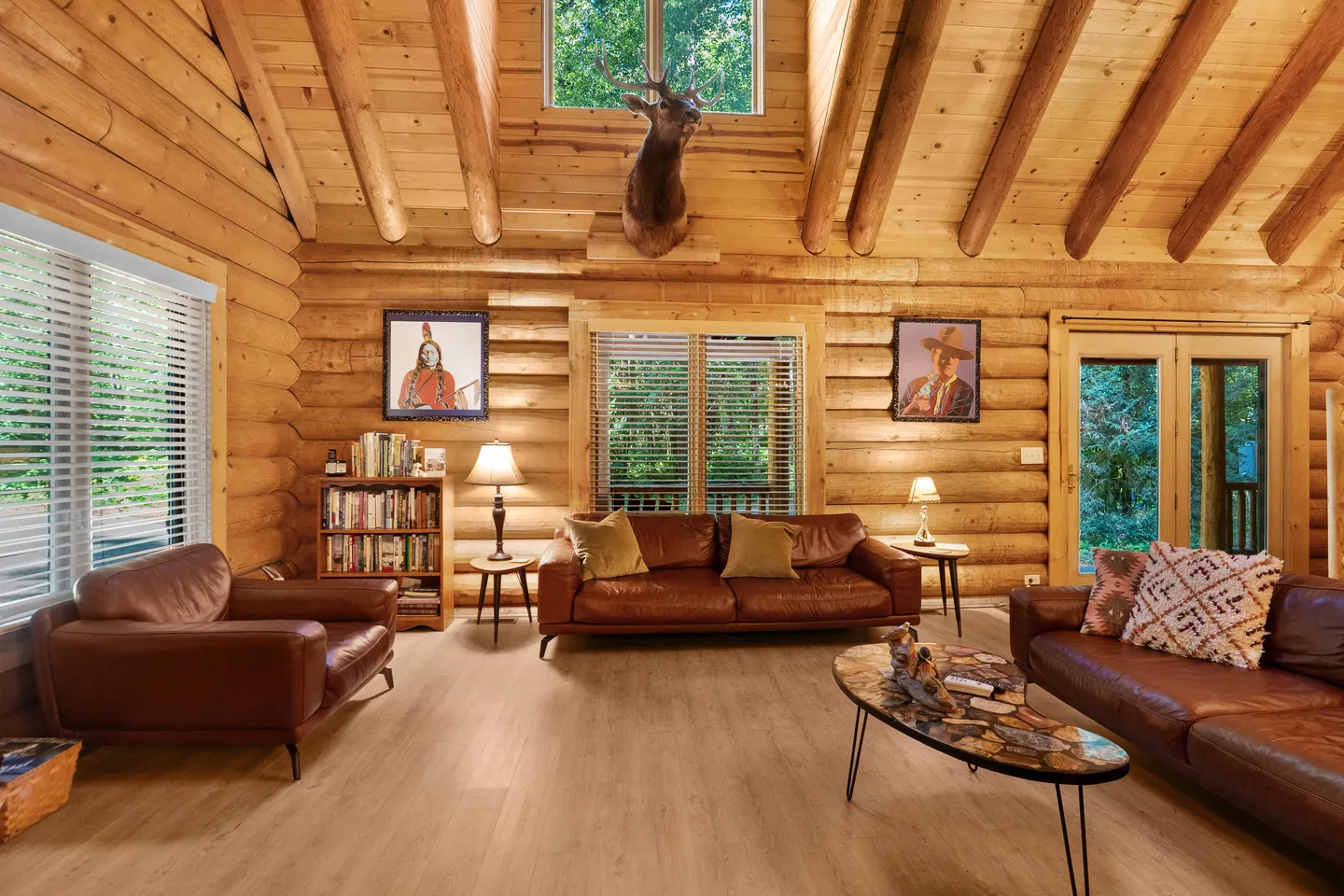 Living room with log walls, vaulted exposed-beam ceiling, leather sofas, an armchair, and a mounted deer head.