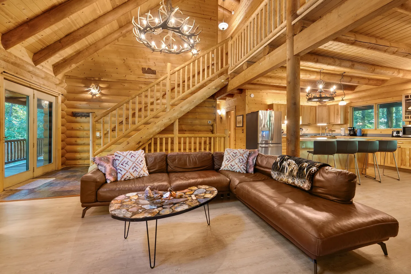 Open living area of a log cabin with a brown leather sectional, stone coffee table, wooden staircase, antler chandelier, and adjacent kitchen.
