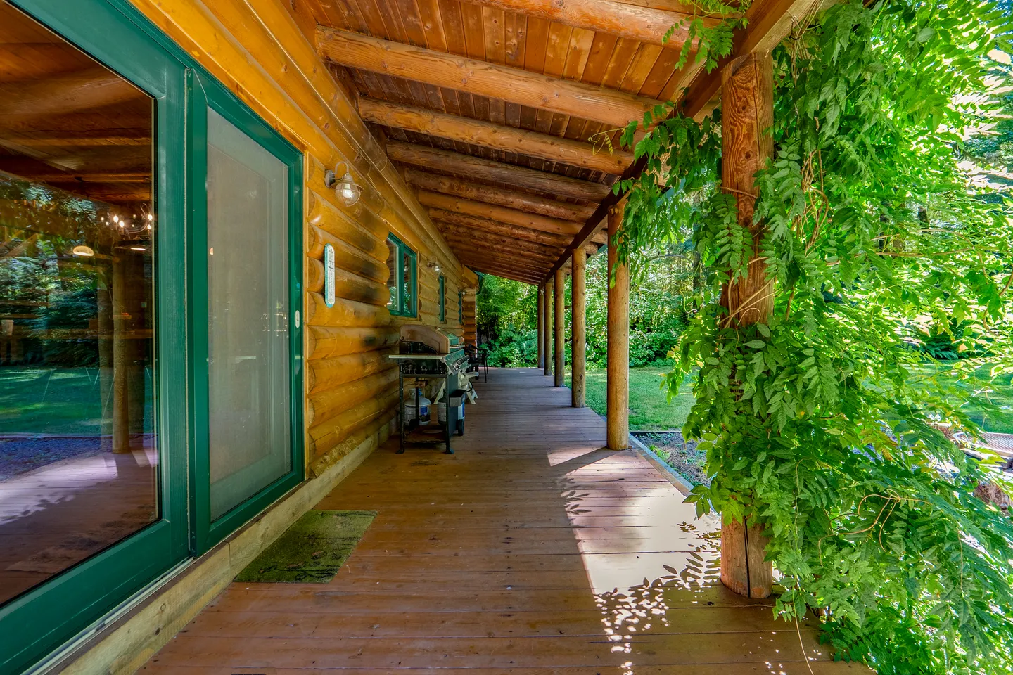 A long covered wooden porch along the log exterior of a cabin, lined with support pillars and lush green foliage.