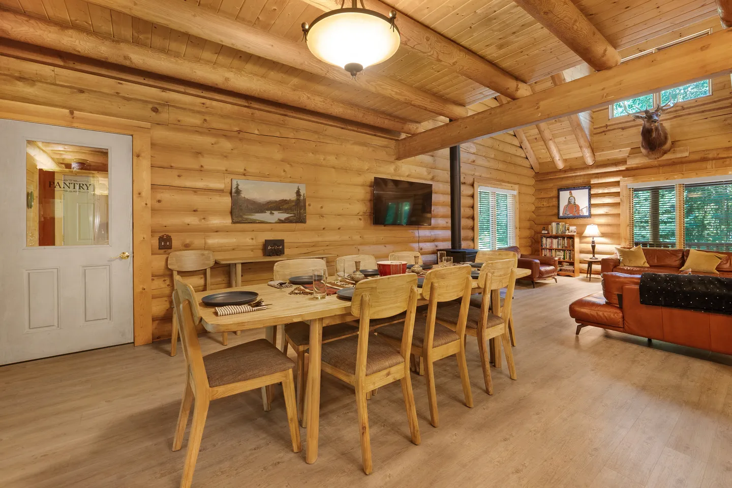 Dining area with a large wooden table and chairs inside a log cabin, adjacent to a living room with leather furniture.