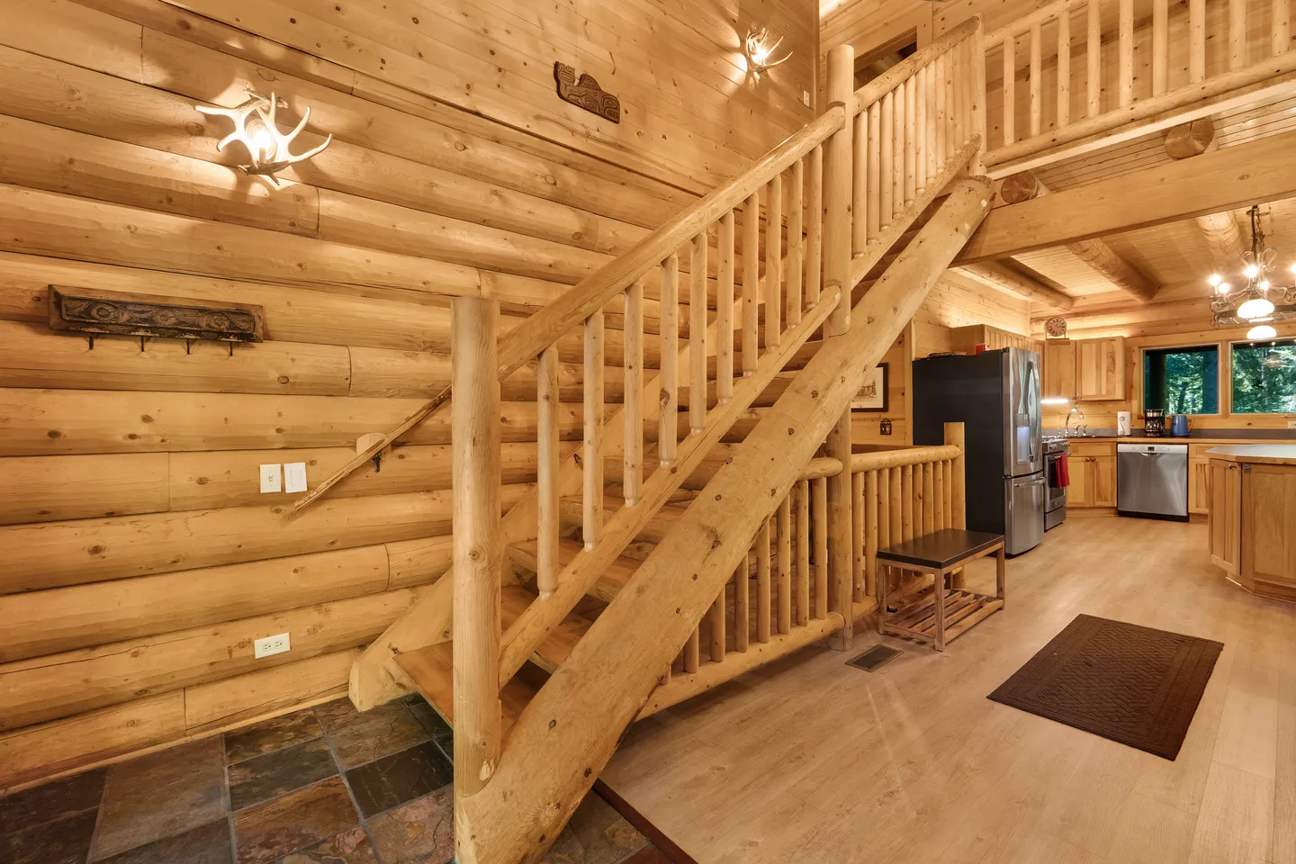 A rustic log cabin interior showing a log-post staircase leading to an upper loft, log walls with antler sconces, and a glimpse of the kitchen.
