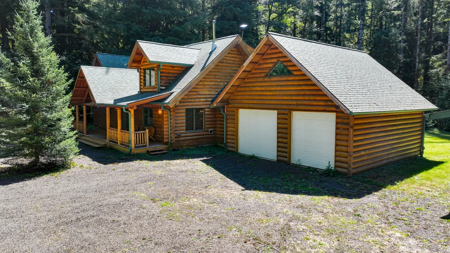 Exterior view of a log cabin home with a covered front porch and an attached two-car garage set against dense woods.