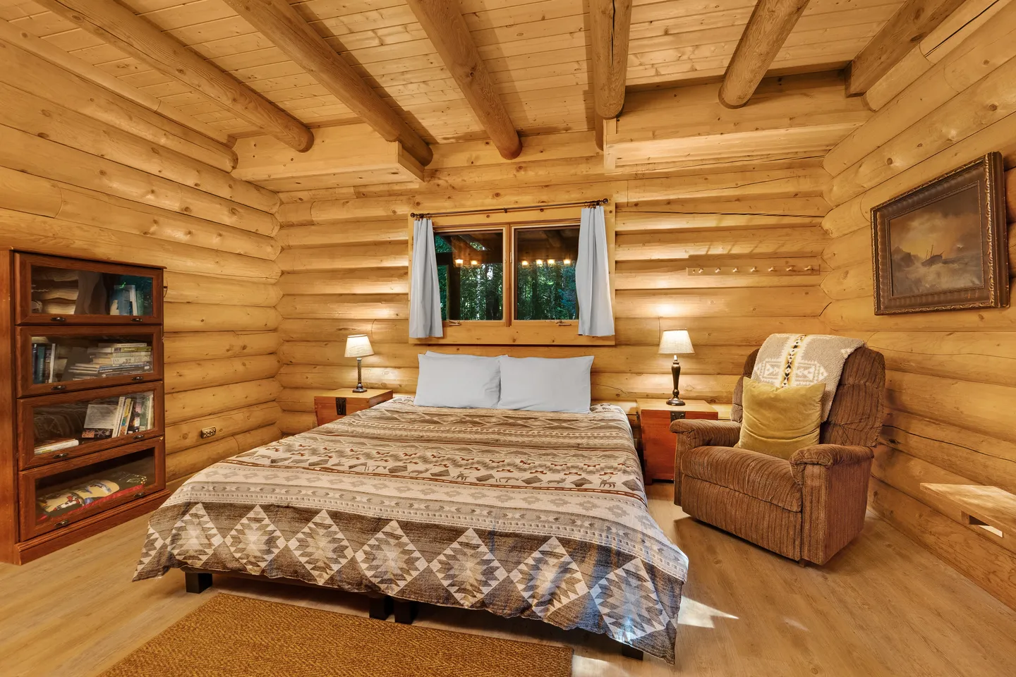 Log cabin bedroom featuring a bed with patterned bedding, flanked by nightstands with lamps, an armchair, and a bookshelf.