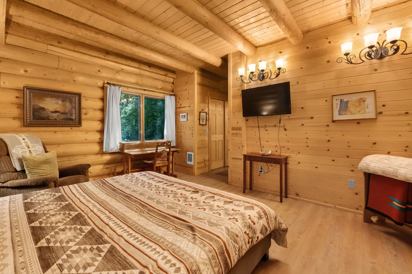Log cabin bedroom with a bed covered in a patterned quilt, wooden walls, a wall-mounted TV, and a small desk by the window.
