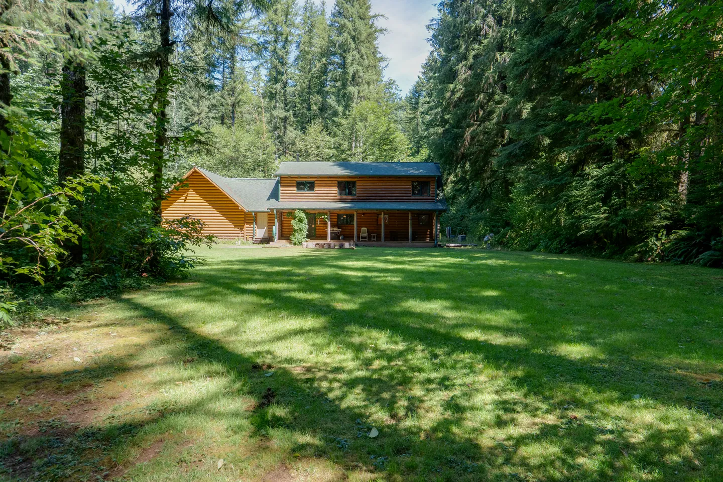 Exterior view of a log cabin nestled in a dense evergreen forest with a large, sun-dappled grass lawn in the foreground.