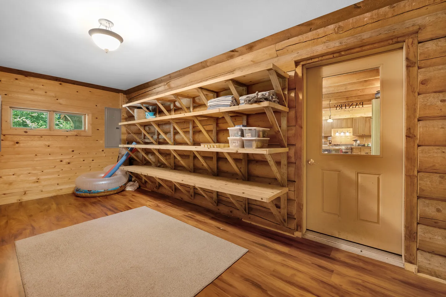 A storage or mudroom area with log siding walls, wide wooden wall-mounted shelves, and a door labeled Pantry.