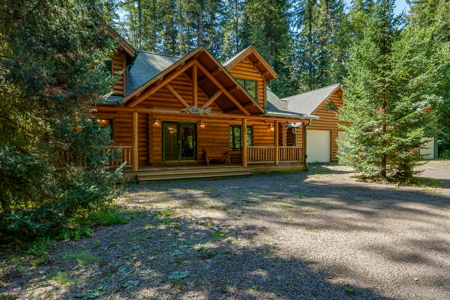 Exterior of a two-story log cabin home with a front porch, gabled rooflines, and an attached garage surrounded by tall pine trees.