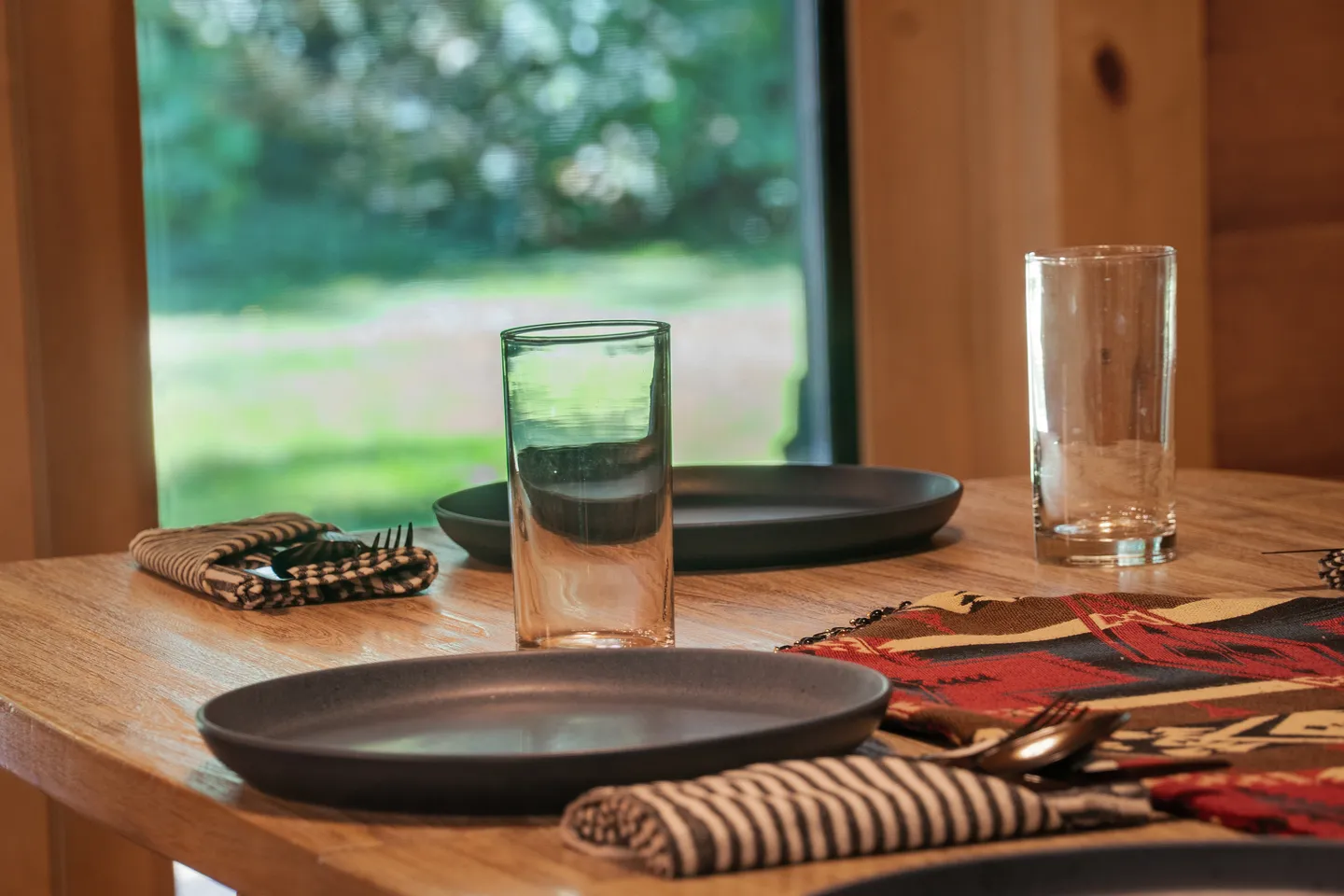 Close-up of a wooden dining table set with dark plates, drinking glasses, and napkins next to a window.