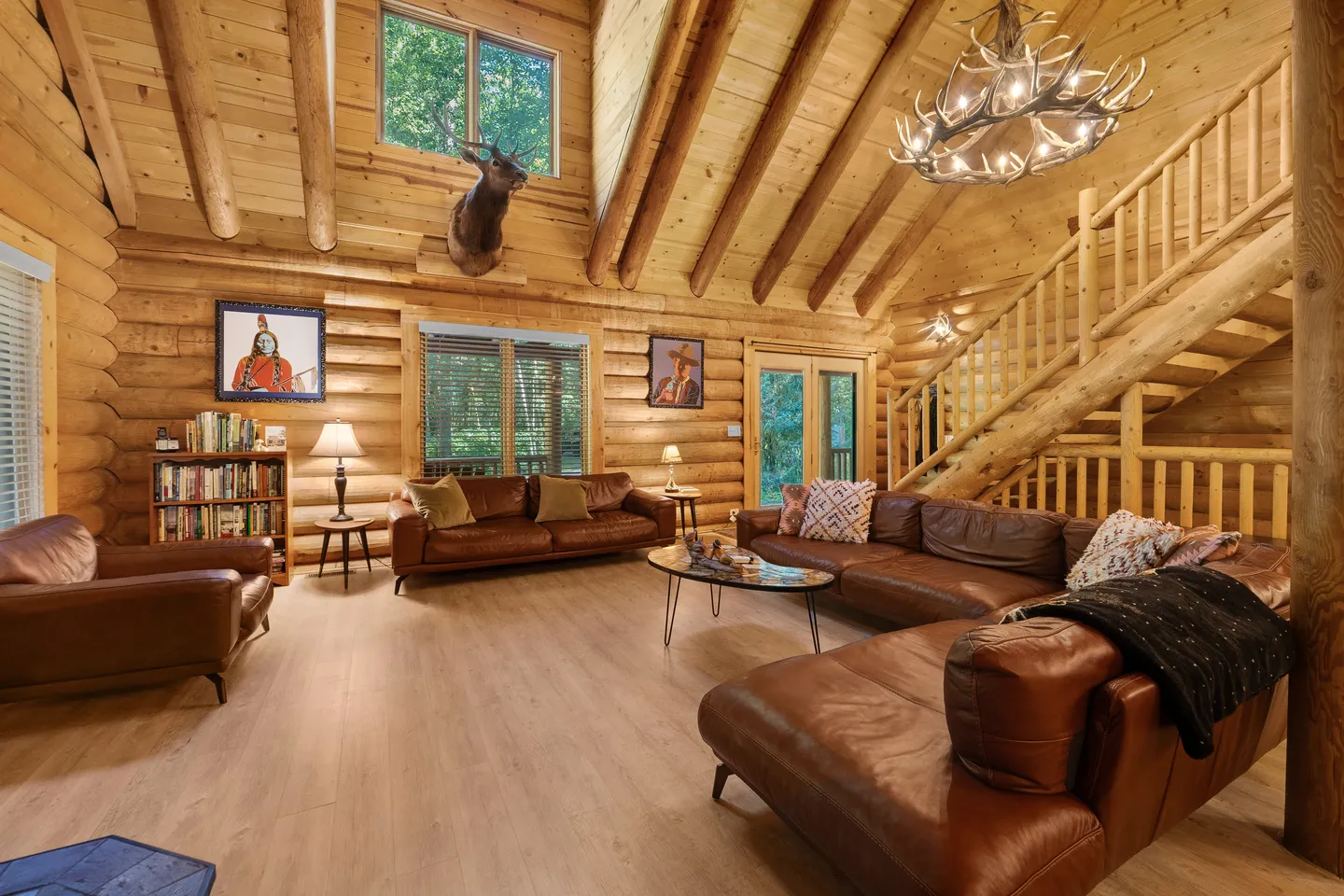Living room with log walls, vaulted wood ceiling, leather sofas, an antler chandelier, and a log staircase.