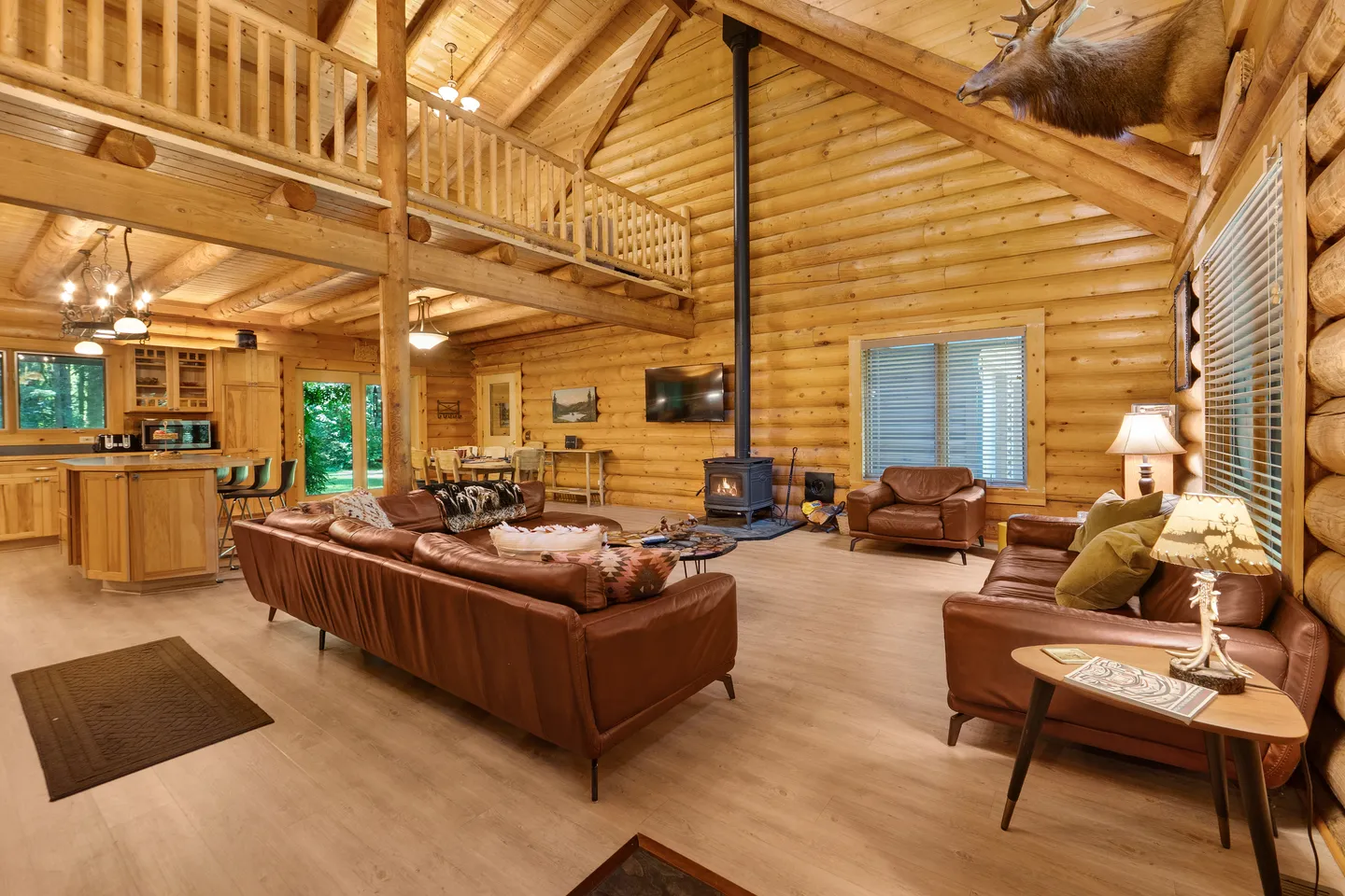 Open-concept log cabin living room and kitchen featuring high vaulted ceilings, a wood stove, leather sofas, and an open loft.