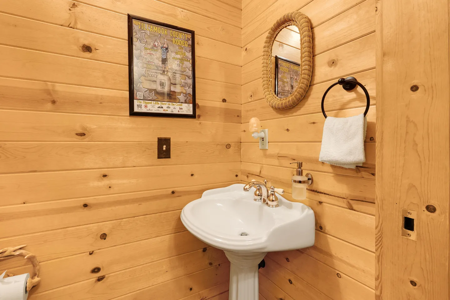 Powder room corner featuring light wood-paneled walls, a white pedestal sink, an oval mirror with a textured rope frame, and a framed rodeo poster.