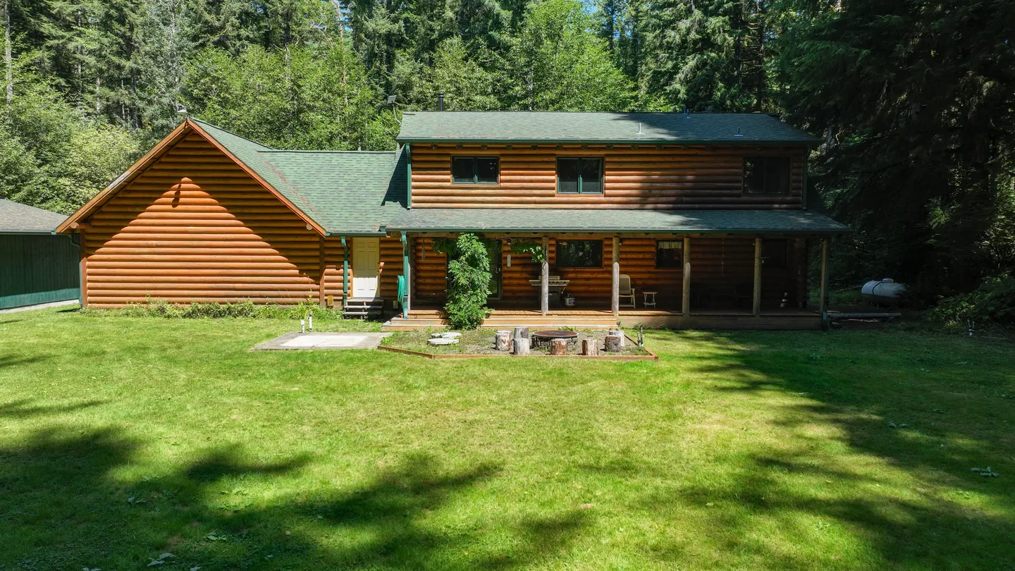 Exterior rear view of a two-story log cabin home with a green roof, covered back porch, and large grassy yard surrounded by trees.