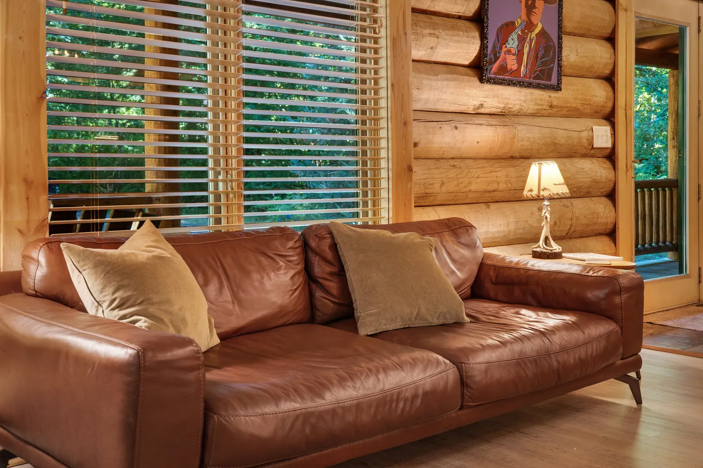 Brown leather sofa with throw pillows in front of a window with blinds inside a log cabin room.