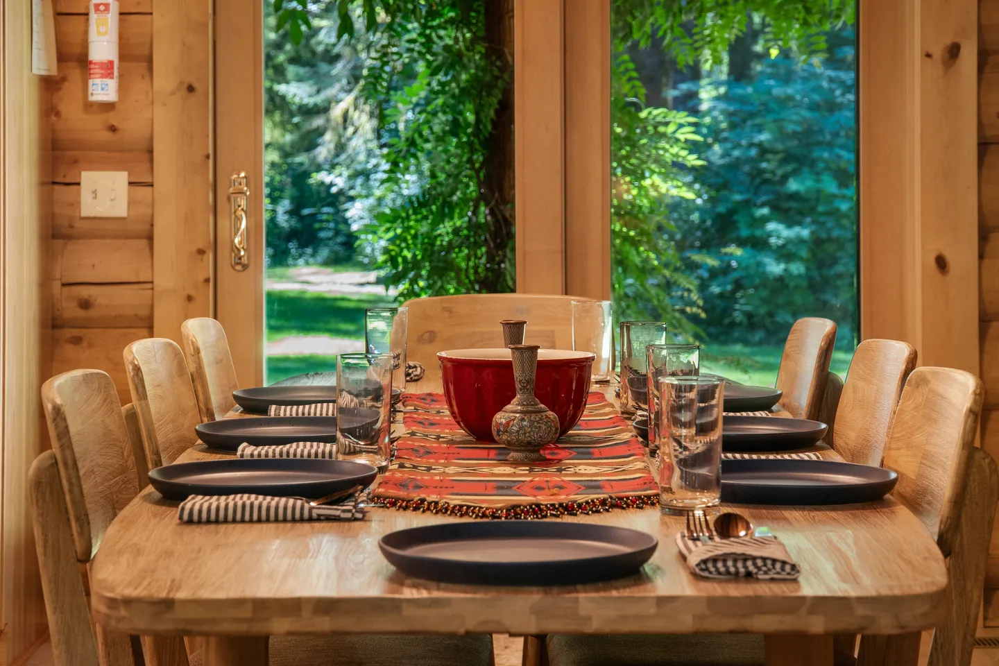 A set wooden dining table lined with chairs facing large glass doors that look out onto lush green trees.