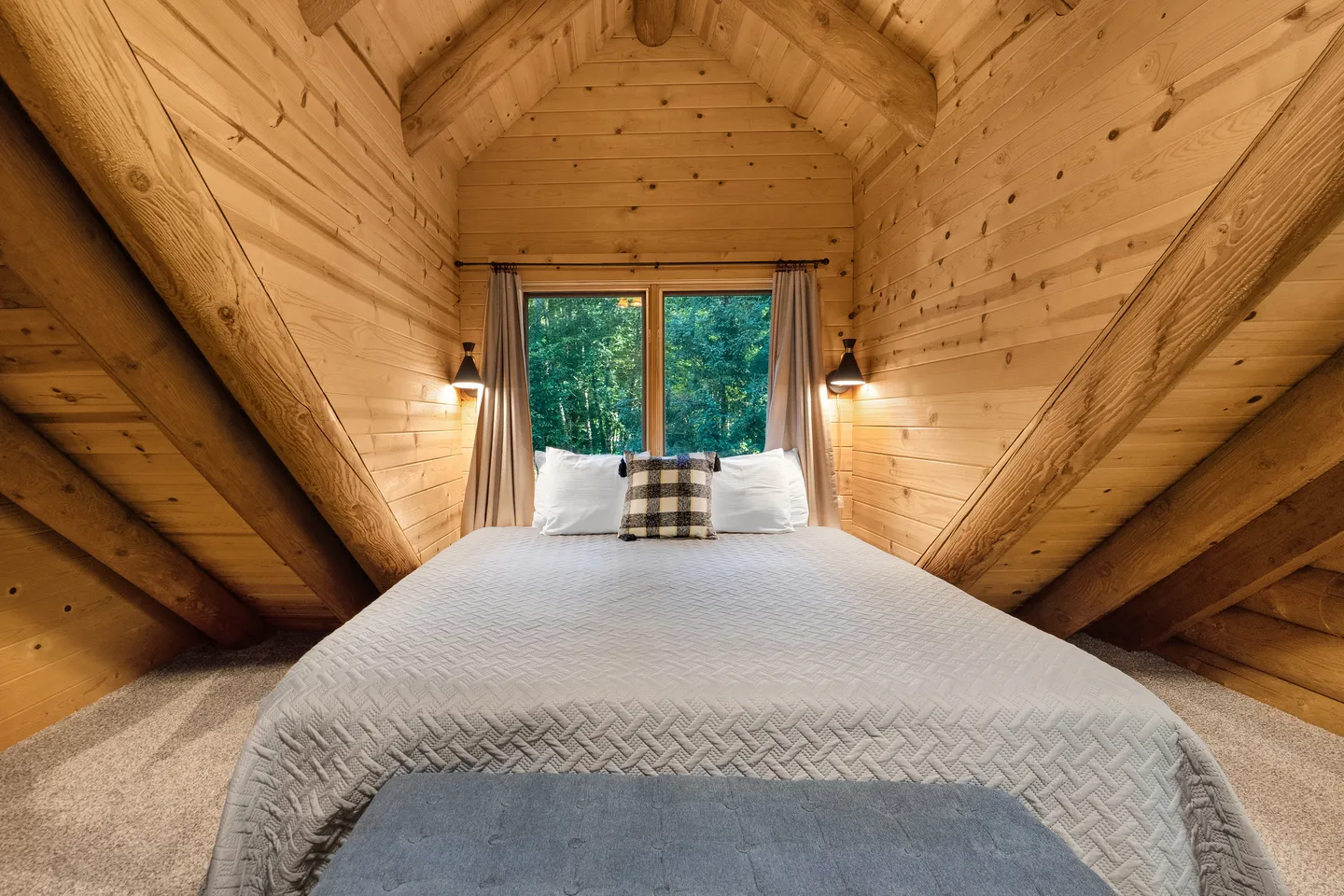 A bed nestled under an A-frame wood-paneled ceiling with a window overlooking green trees.