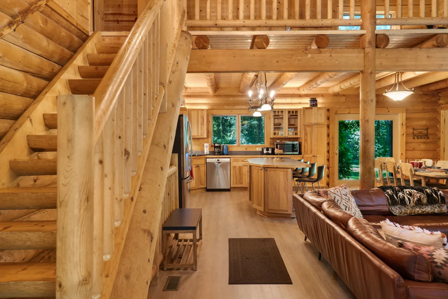 Open-concept log cabin interior showing a wooden staircase, living area with leather couch, kitchen, and dining space.
