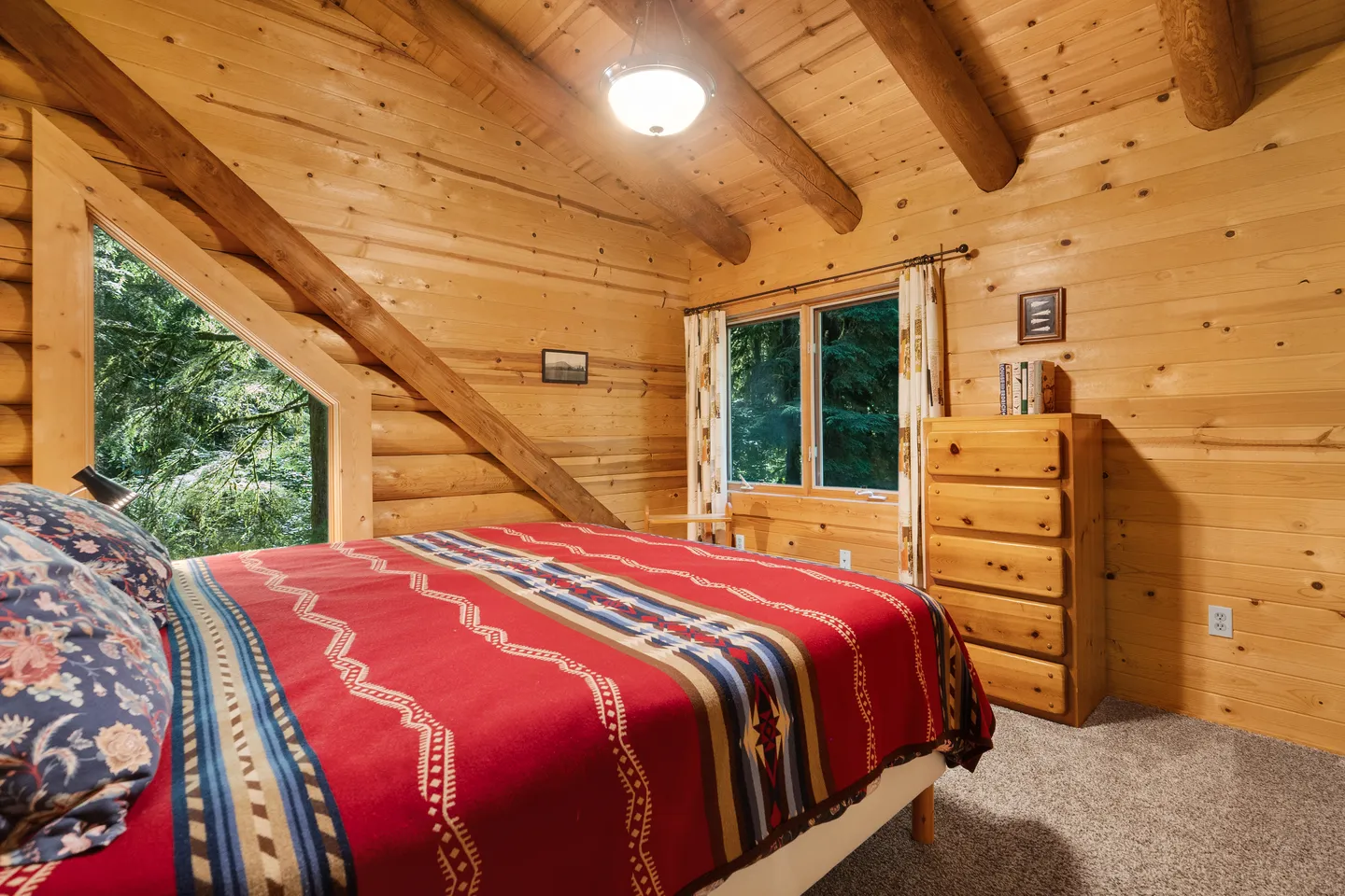 Bedroom with log cabin wood walls, exposed log beams, a patterned red bedspread, and windows overlooking trees.