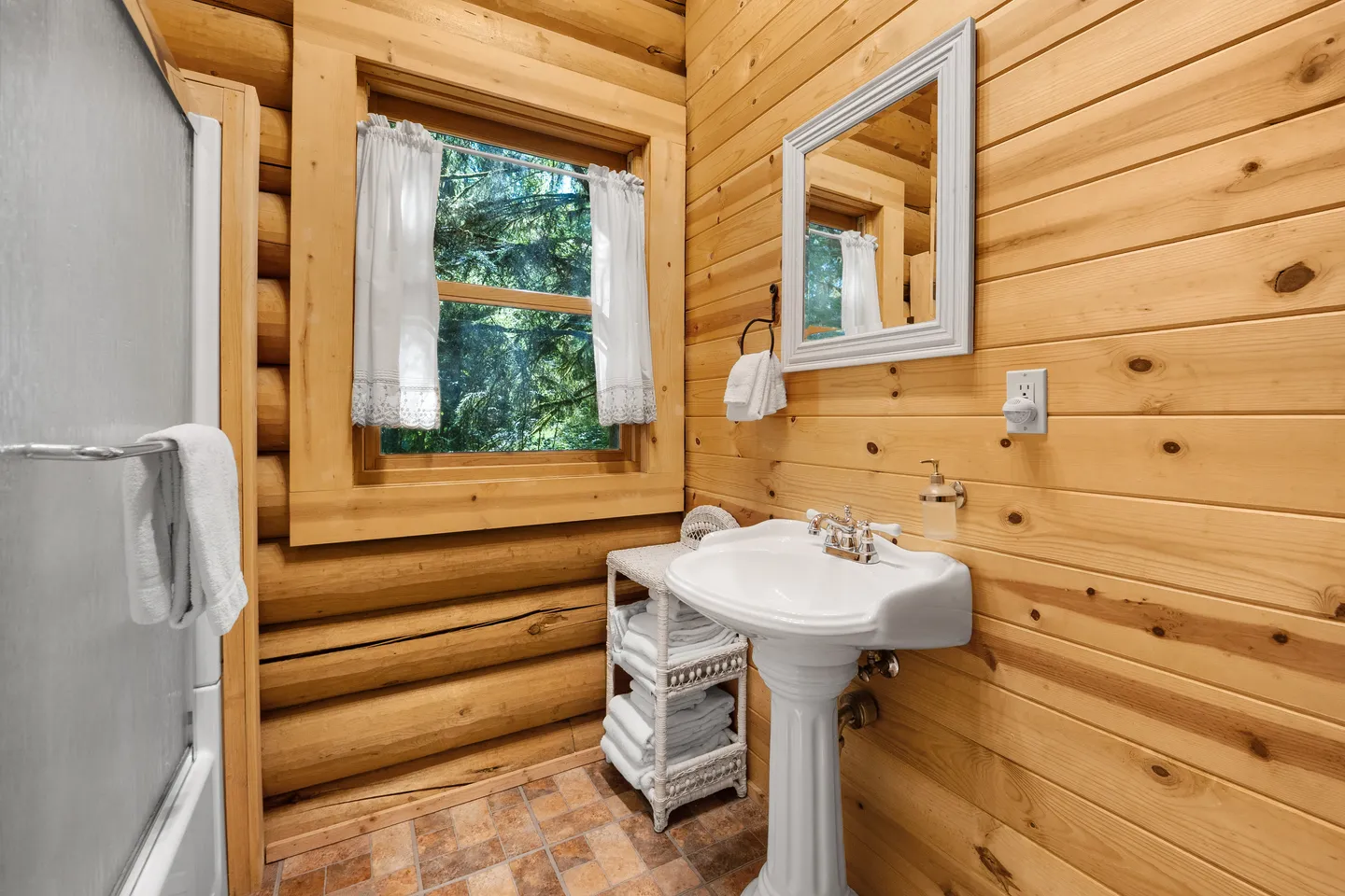 Bathroom with log-paneled walls, a white pedestal sink, white-framed mirror, and a window with white lace curtains.