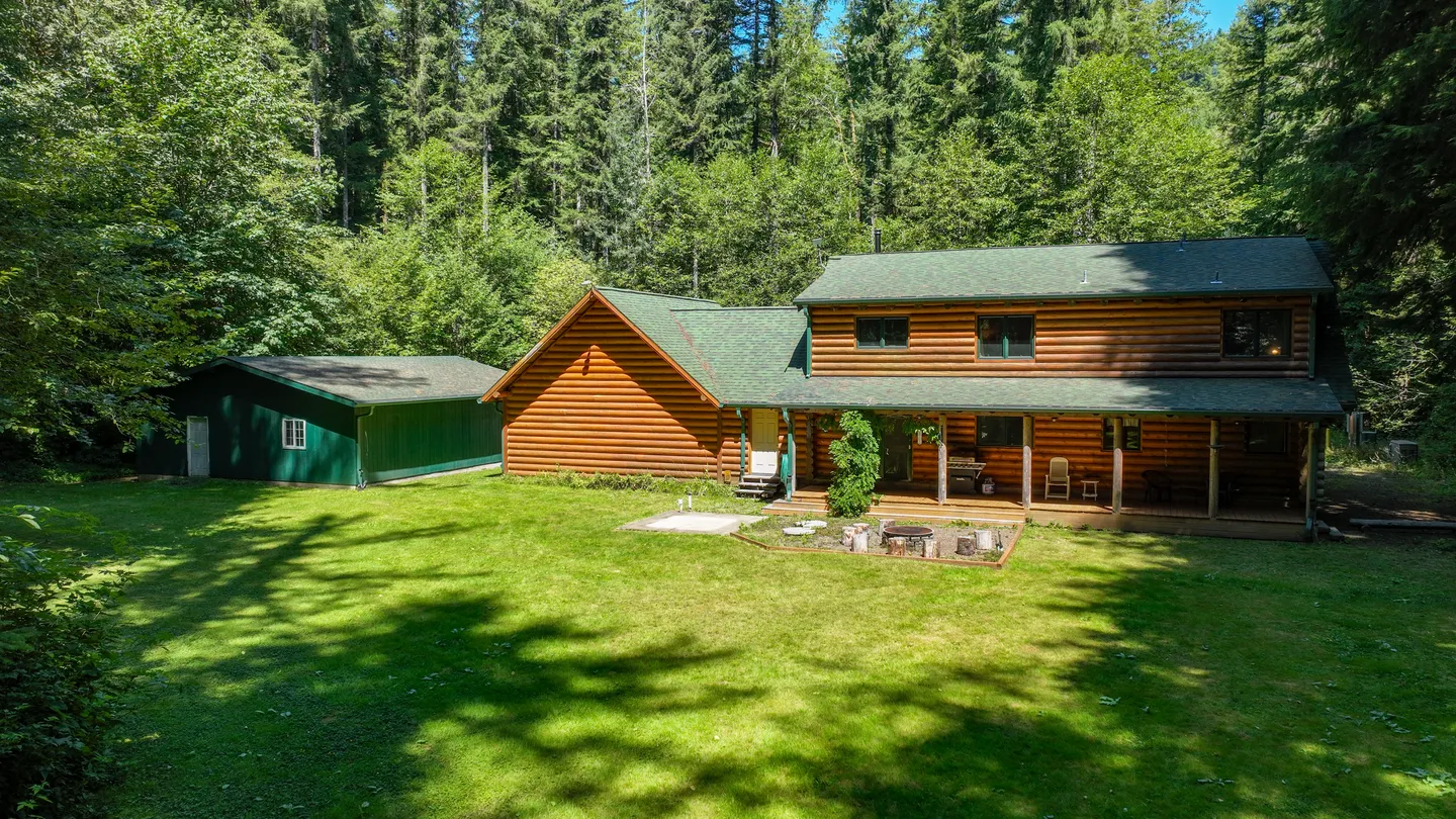 Exterior view of a two-story log cabin with a covered back porch, adjacent green outbuilding, and a large grassy yard bordered by tall evergreen trees.