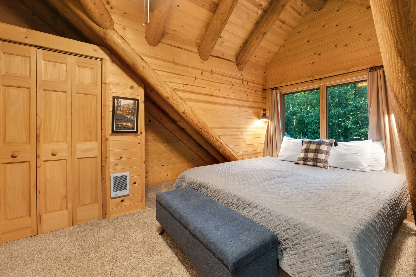 Rustic log cabin bedroom with a bed beneath a vaulted beamed ceiling, closet, and window showing greenery.