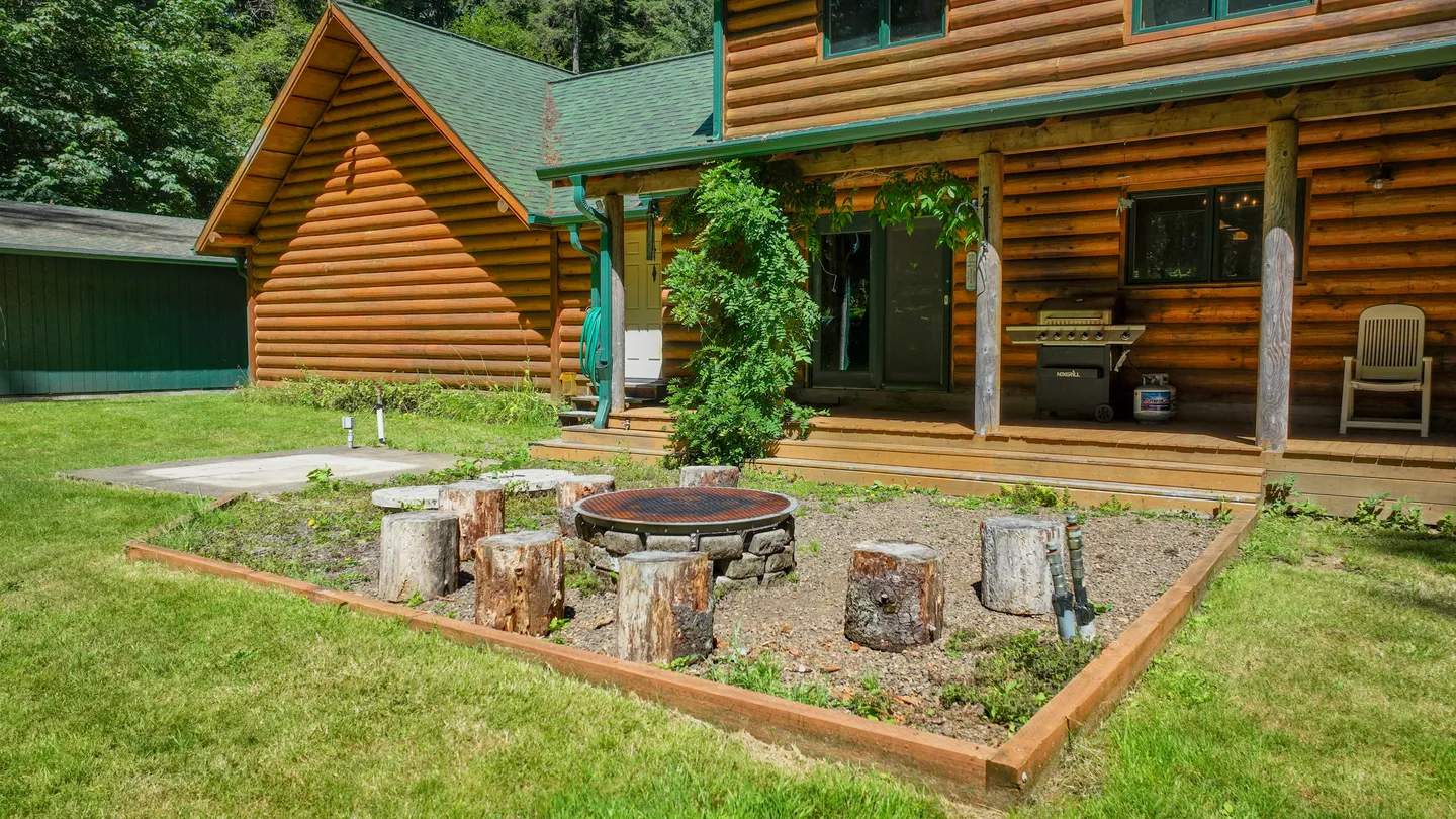 Outdoor fire pit area with tree stump seating in front of a log cabin with a covered back porch.