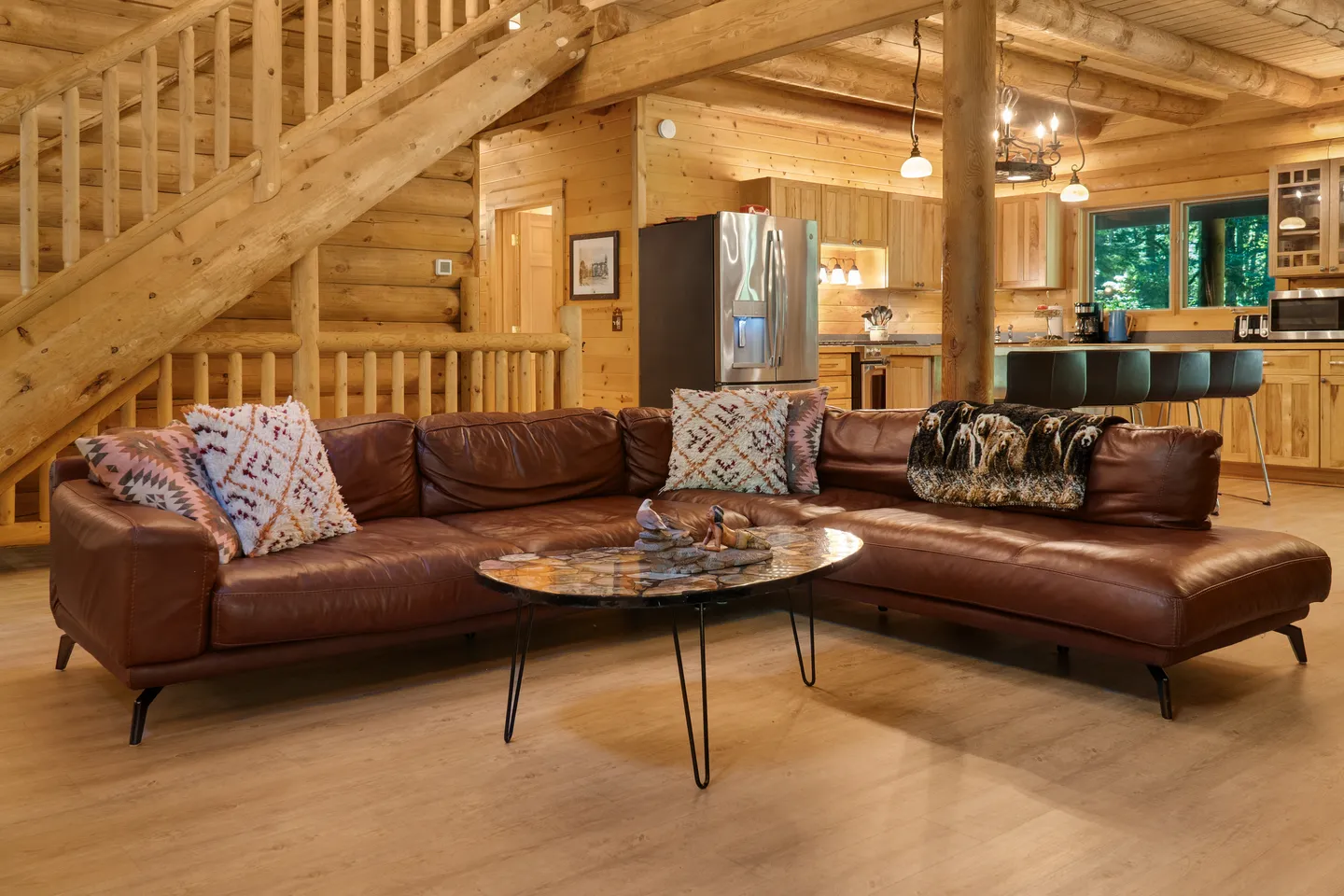 Open-concept log cabin living area with a brown leather sectional, coffee table, wood staircase, and kitchen in the background.