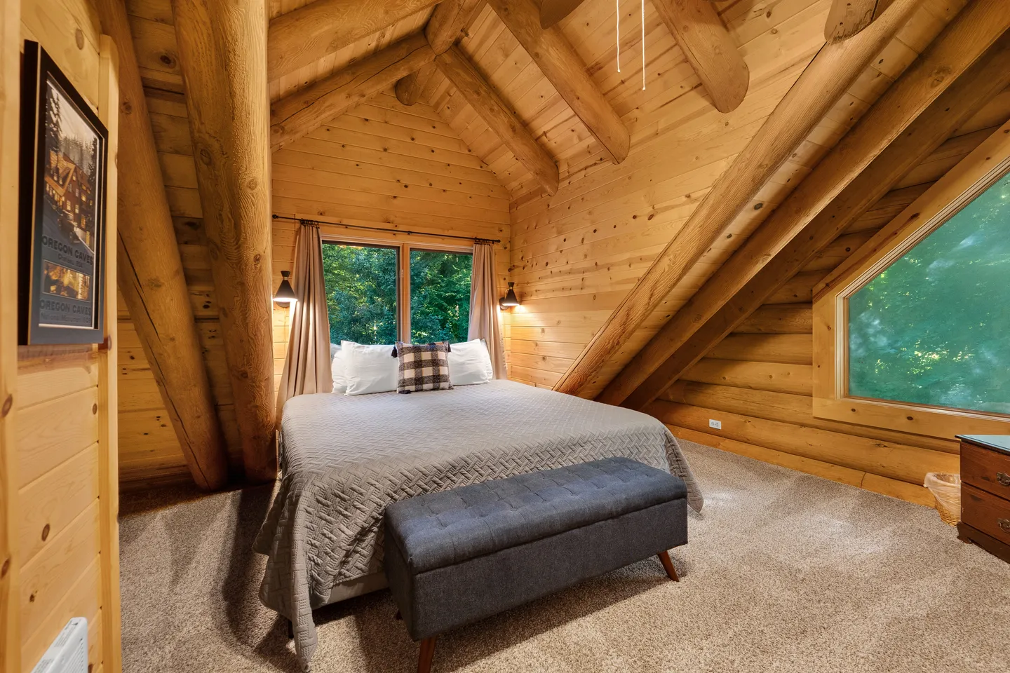 Loft bedroom with log cabin walls, vaulted ceiling with heavy log beams, carpeted floor, bed, and upholstered bench.