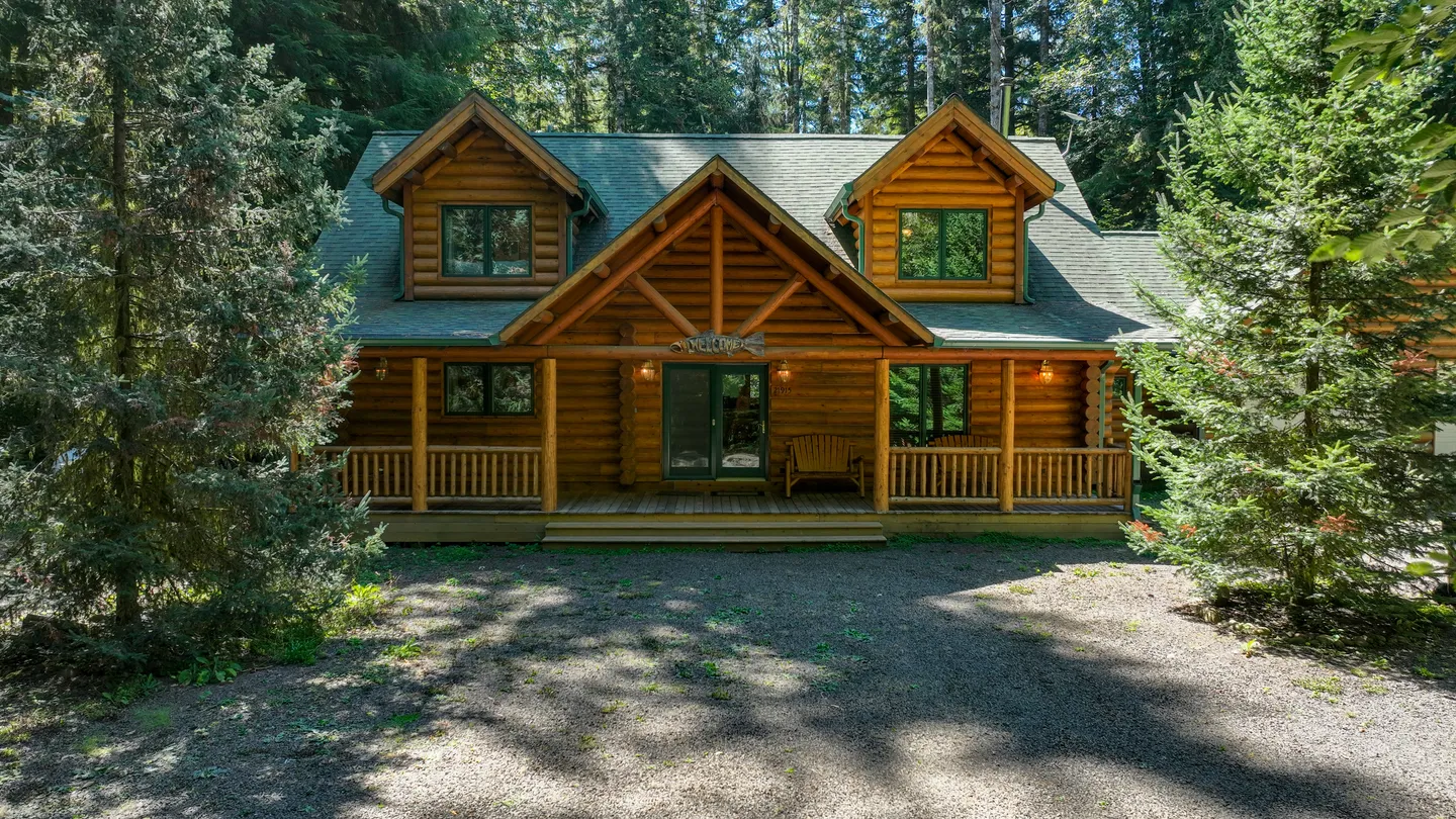 Exterior view of a log cabin with a covered front porch surrounded by tall evergreen trees.