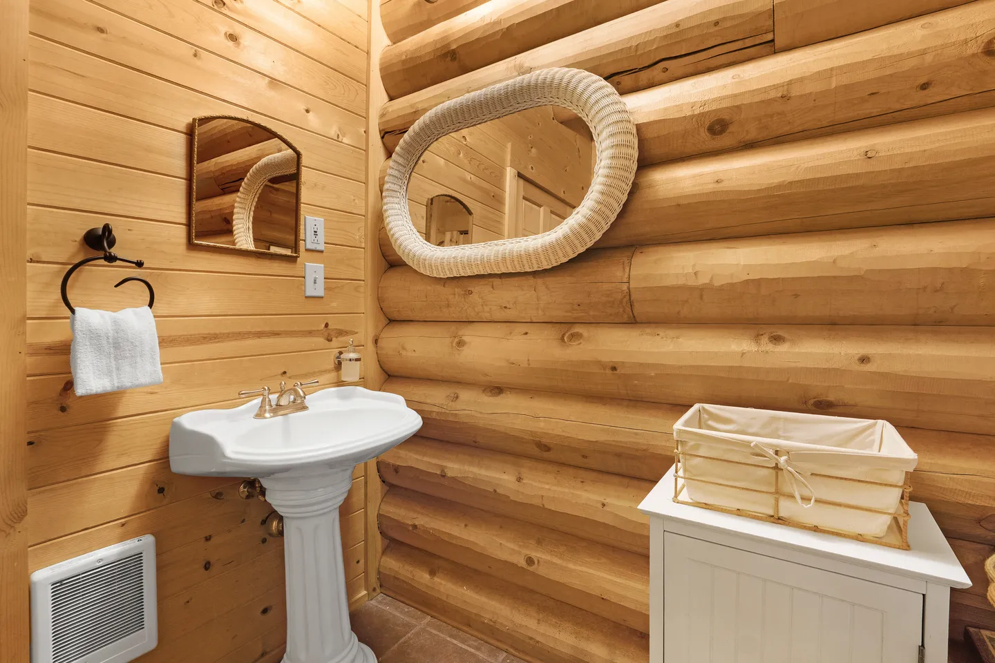 Bathroom with log-cabin walls, a white pedestal sink, a wicker-framed mirror, and a small storage cabinet.
