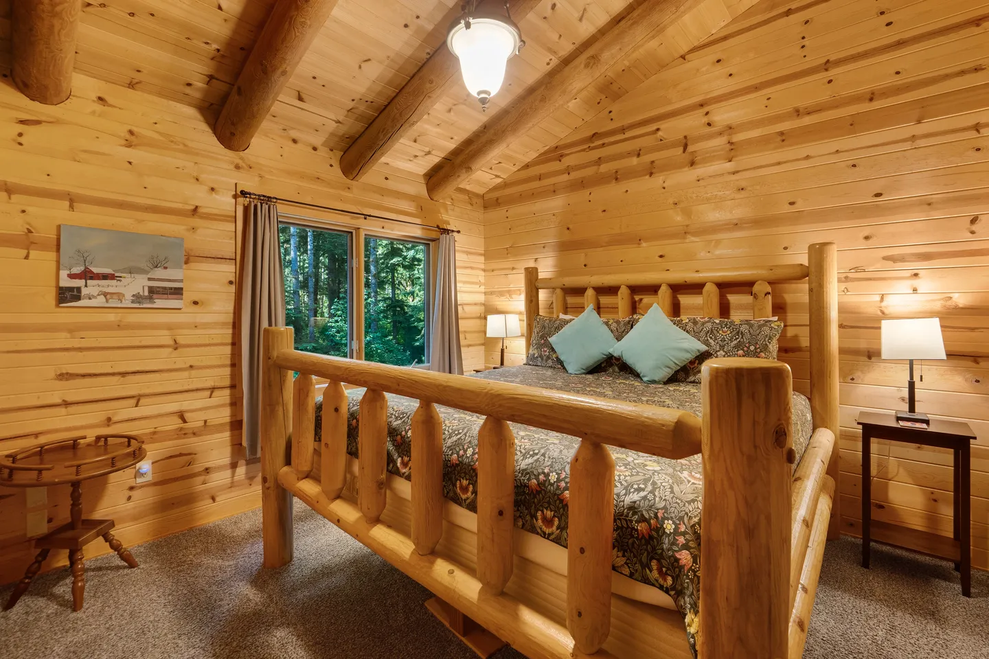 Bedroom with log frame bed, wood-paneled walls and vaulted log beam ceiling, carpeted flooring, and a window viewing green trees.