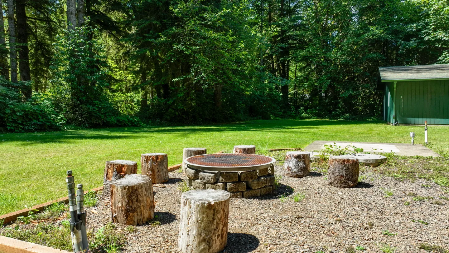 Outdoor fire pit with a metal grate surrounded by tree stump seats in a gravel area next to a grassy lawn and forest.