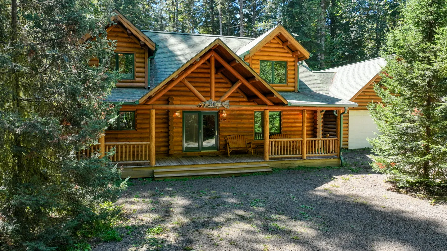 Exterior of a two-story log cabin surrounded by trees, featuring a covered front porch and a gravel driveway.