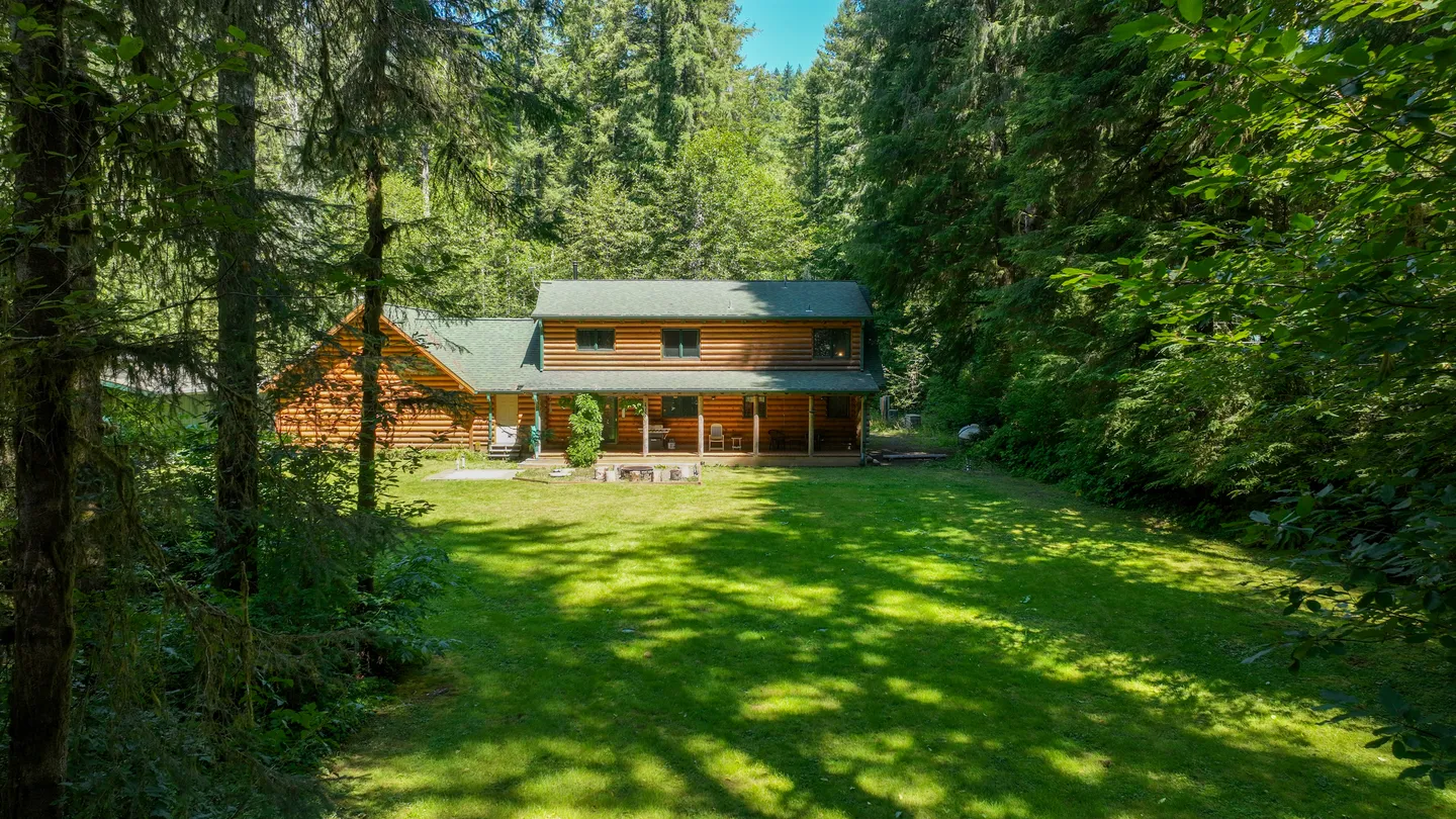 A two-story log cabin house viewed across a spacious green lawn, surrounded by dense evergreen trees.