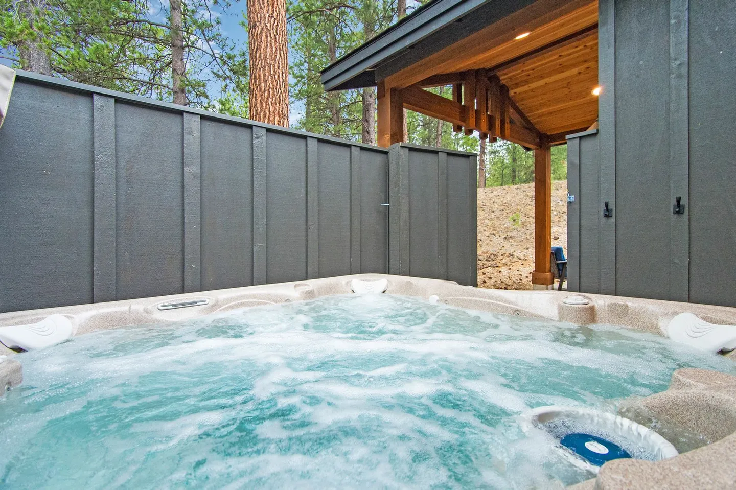 Close-up view of an outdoor hot tub with bubbling water next to a dark grey privacy fence and a covered structure surrounded by tall trees.