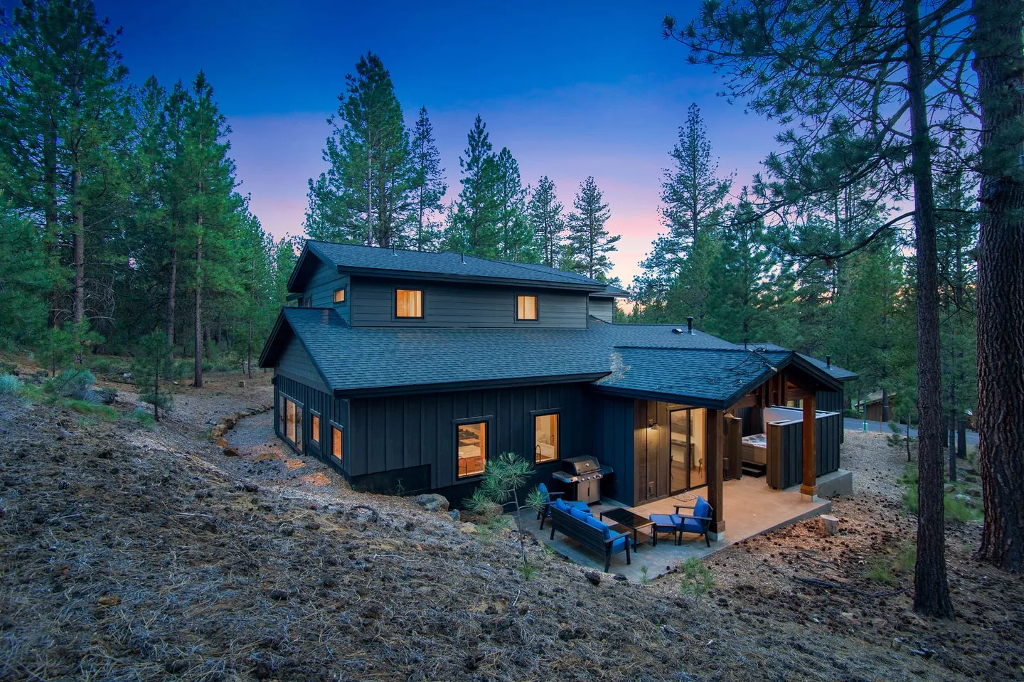 Exterior dusk view of a modern dark-sided home nestled in a pine forest with a patio and hot tub.