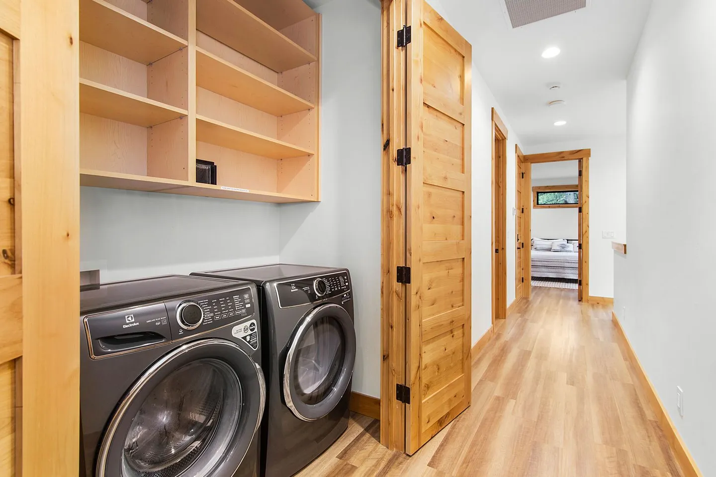 Hallway laundry closet with front-loading washer and dryer, open wooden shelves, and bi-fold wooden doors.