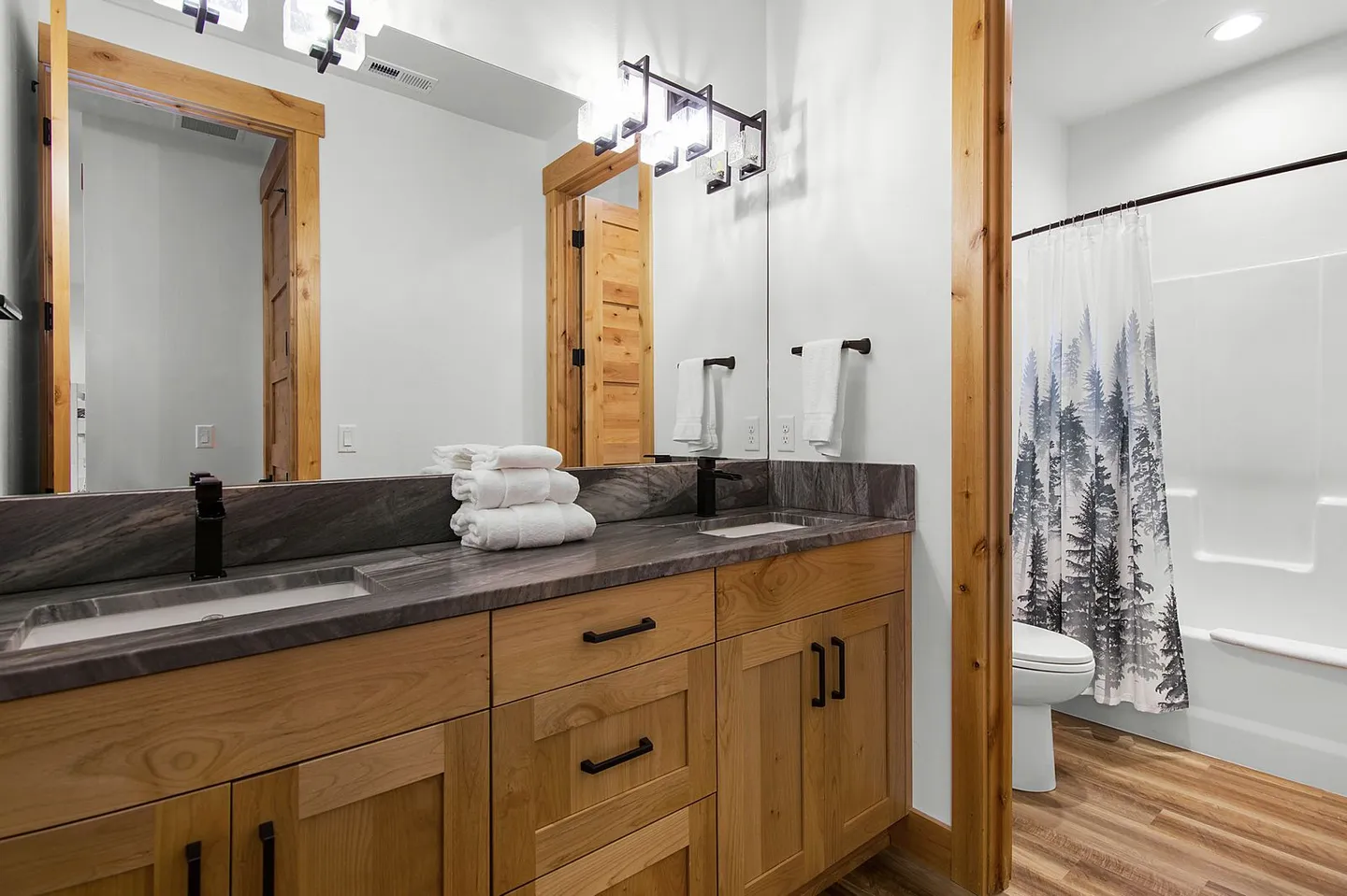 Bathroom with a double sink wooden vanity, dark countertops, large mirror, and a view into the toilet and shower area.