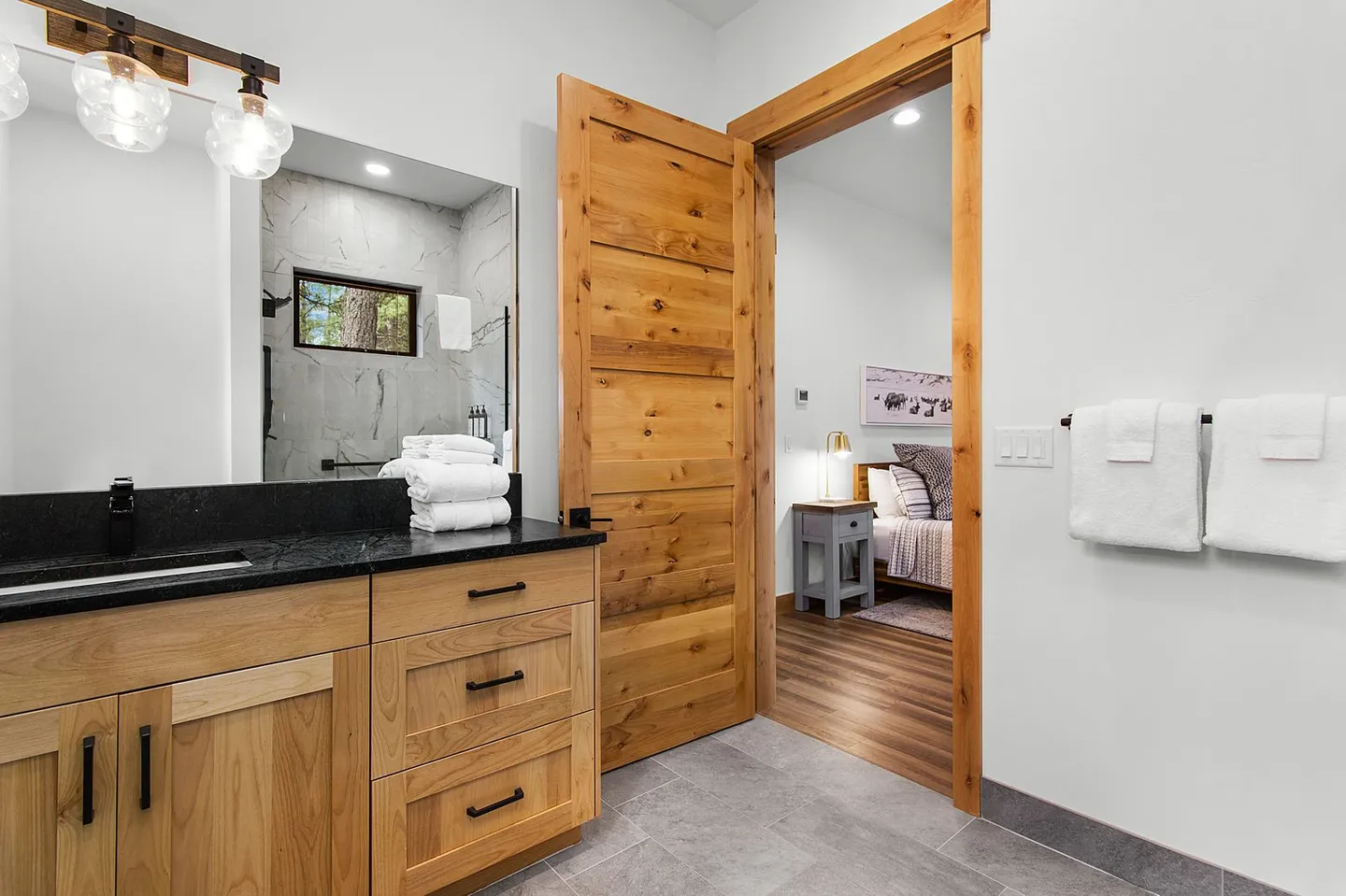Bathroom with light wood vanity, dark countertop, and an open wood-paneled door leading into a bedroom.