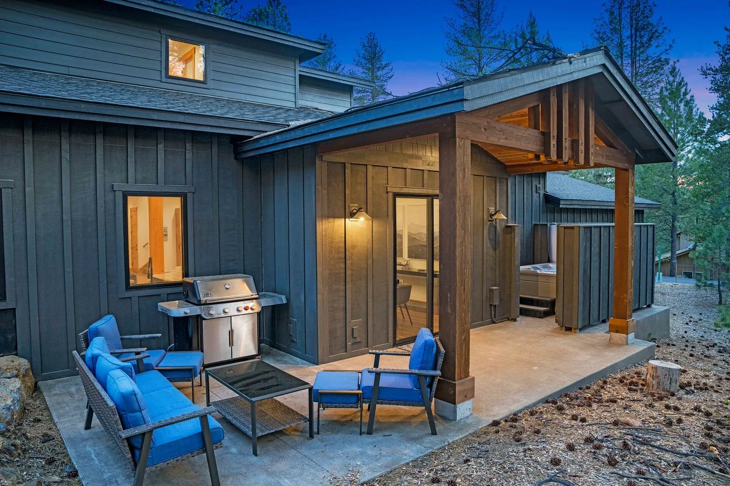 Outdoor concrete patio at dusk featuring dark vertical siding, a covered porch section, blue cushioned patio seating, a gas barbecue grill, and a hot tub tucked into a recessed nook.