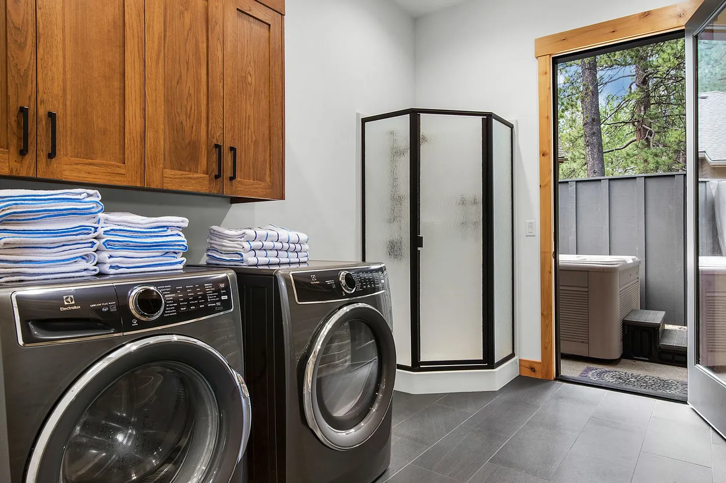 A laundry room with front-loading washer and dryer, upper wooden cabinets, a corner walk-in shower, and an open exterior door leading to a patio with a hot tub.