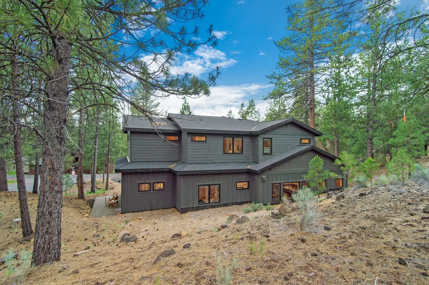 Exterior of a two-story dark gray house nestled on a rocky, pine-covered hillside under a partly cloudy sky.