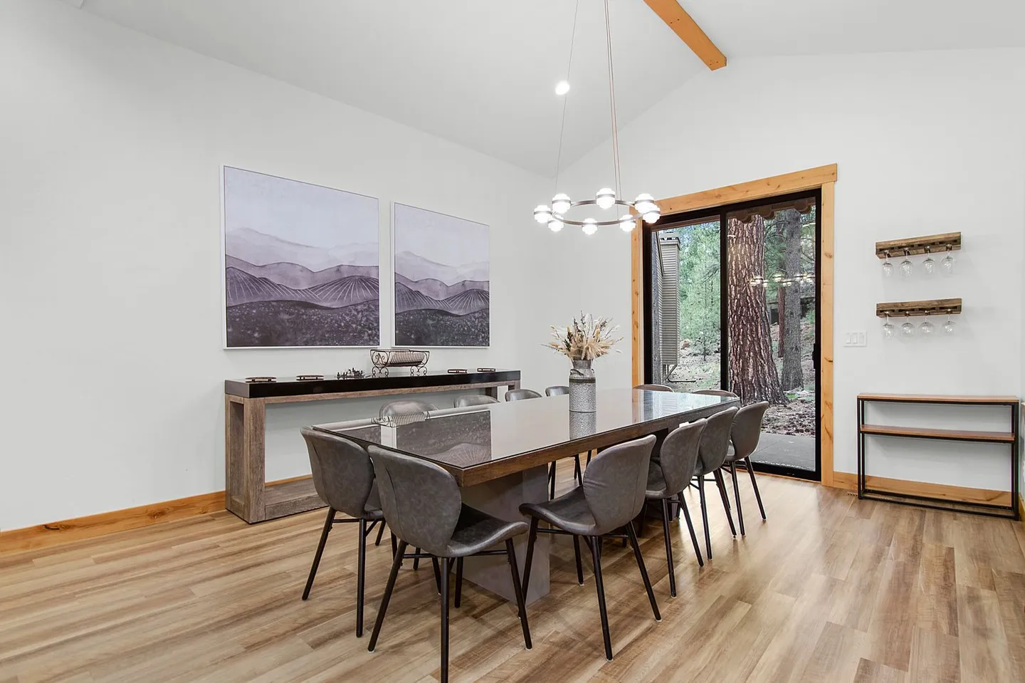 Dining room with a long table, grey upholstered chairs, modern ring chandelier, and sliding glass door looking out to trees.