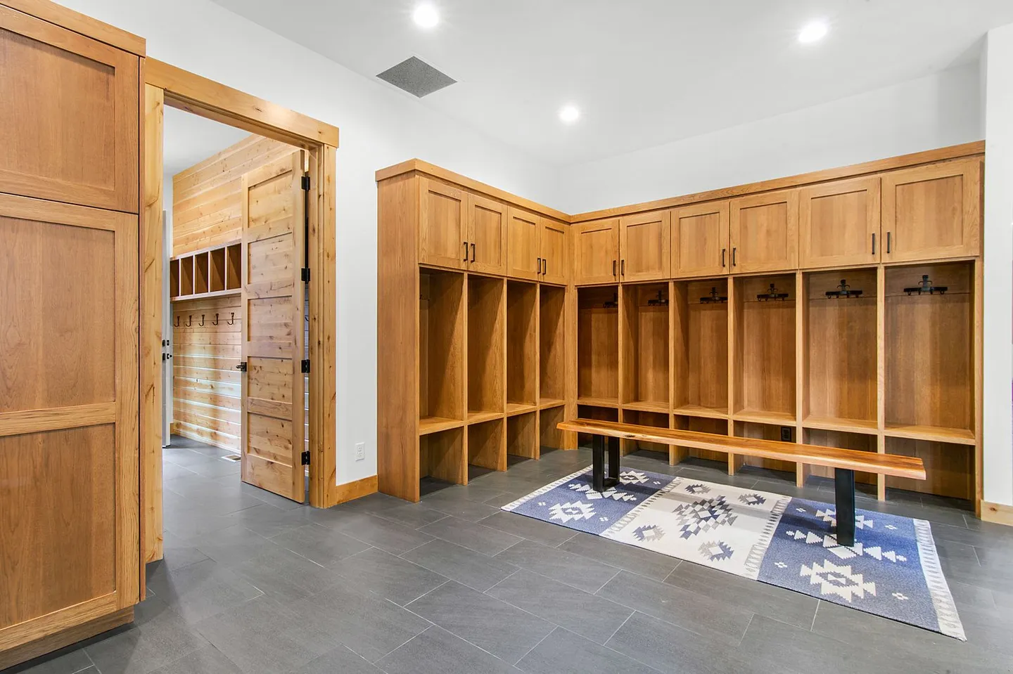 Mudroom or ski locker room featuring custom built-in wood lockers, a wooden bench, patterned area rug, and dark tile flooring.