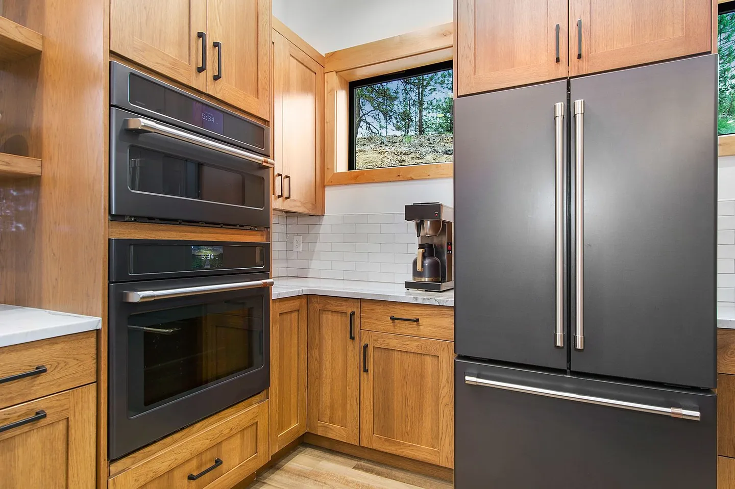 Kitchen corner showing built-in wall ovens, matte black refrigerator, light wood cabinetry, and a coffee maker on the counter by a window.