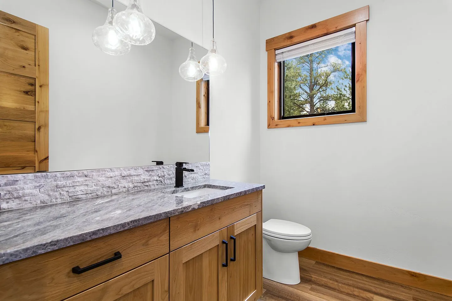 Bathroom vanity with stone-look countertop, wood cabinetry, large mirror, pendant lights, toilet, and a wood-trimmed window.